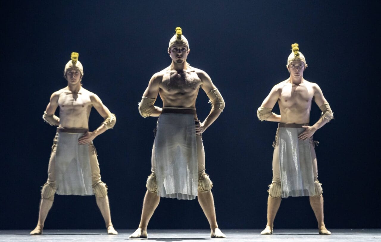 Three male dancers stand on stage in formation, wearing ancient warrior-inspired costumes with helmets, arm bands, and sheer skirts, illuminated by stage lighting reminiscent of the regal court of Mary, Queen of Scots.