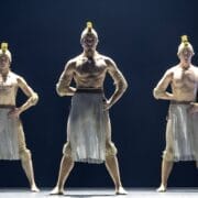 Three male dancers stand on stage in formation, wearing ancient warrior-inspired costumes with helmets, arm bands, and sheer skirts, illuminated by stage lighting reminiscent of the regal court of Mary, Queen of Scots.