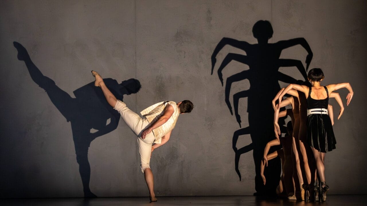A dancer in white kicks with an extended leg while a group in black creates a shadow resembling a spider on a plain wall, evoking the intrigue and drama of Mary Queen of Scots and the Scottish monarchy.