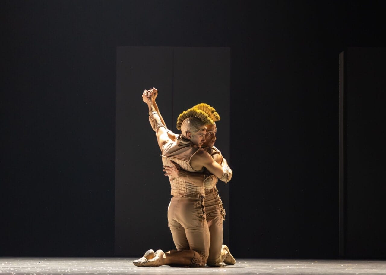 Two performers in beige costumes kneel on stage, embracing each other tightly beneath dramatic lighting and a dark background, evoking the tense intimacy of Mary Queen of Scots within the Scottish monarchy.