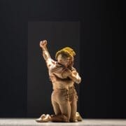 Two performers in beige costumes kneel on stage, embracing each other tightly beneath dramatic lighting and a dark background, evoking the tense intimacy of Mary Queen of Scots within the Scottish monarchy.