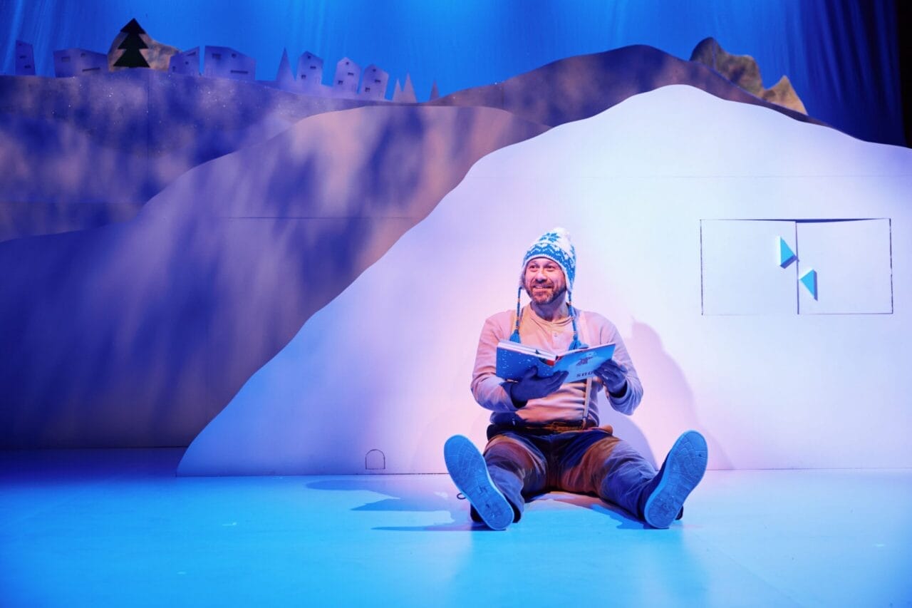 A man in winter clothing sits on the floor in front of a white, snow-like set, reading a book under blue stage lighting.