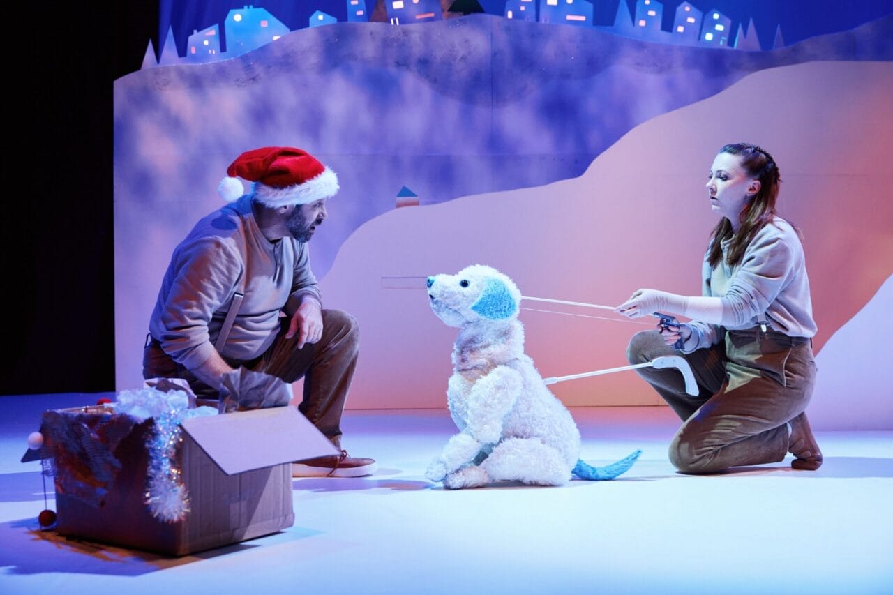 Two people on stage interact with a large polar bear puppet; one wears a Santa hat, and snow-themed scenery is in the background.