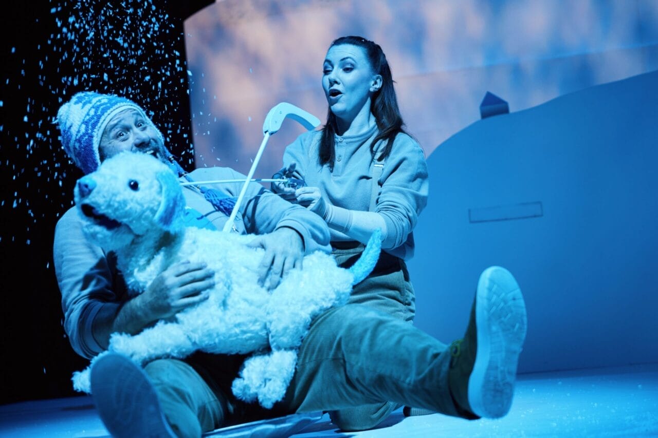 Two actors on stage in winter attire sit on snowy ground, holding a white dog puppet as artificial snow falls around them, under blue stage lighting.