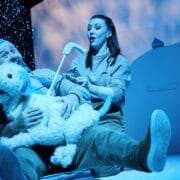 Two actors on stage in winter attire sit on snowy ground, holding a white dog puppet as artificial snow falls around them, under blue stage lighting.