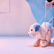 A person controls a large white and blue dog puppet using visible rods on a minimalist stage with pastel colors.