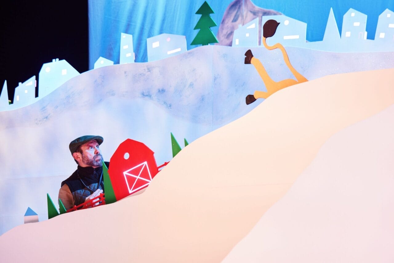 A man in winter clothing looks up from behind a snowy hill set, with a barn prop and the legs of an animal sticking out of the snow. White buildings and trees are in the background.