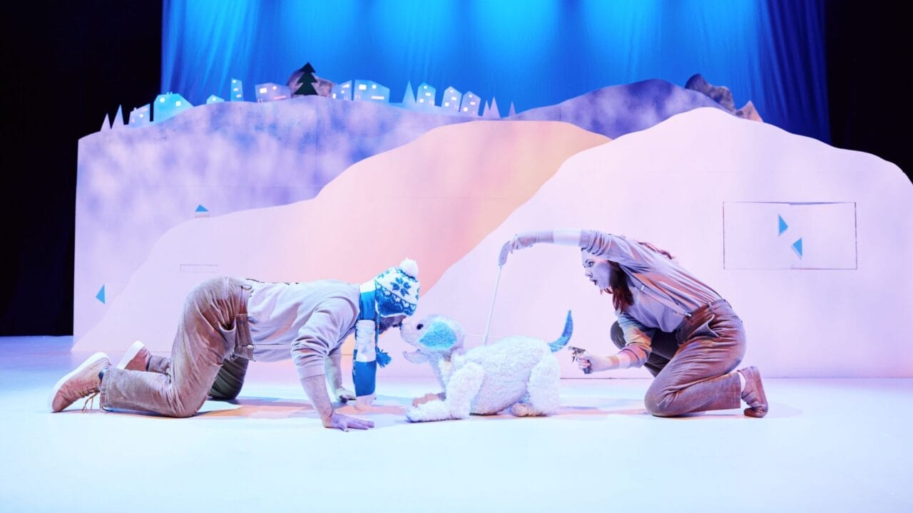 Two performers in costumes kneel and interact with a white dog puppet on a snowy stage set with blue lighting and a backdrop of stylized hills and small houses.