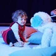 A young child sits on the floor smiling at a white, fluffy dog puppet, while an adult operates the puppet nearby. Pillows and a striped cushion are on the floor.