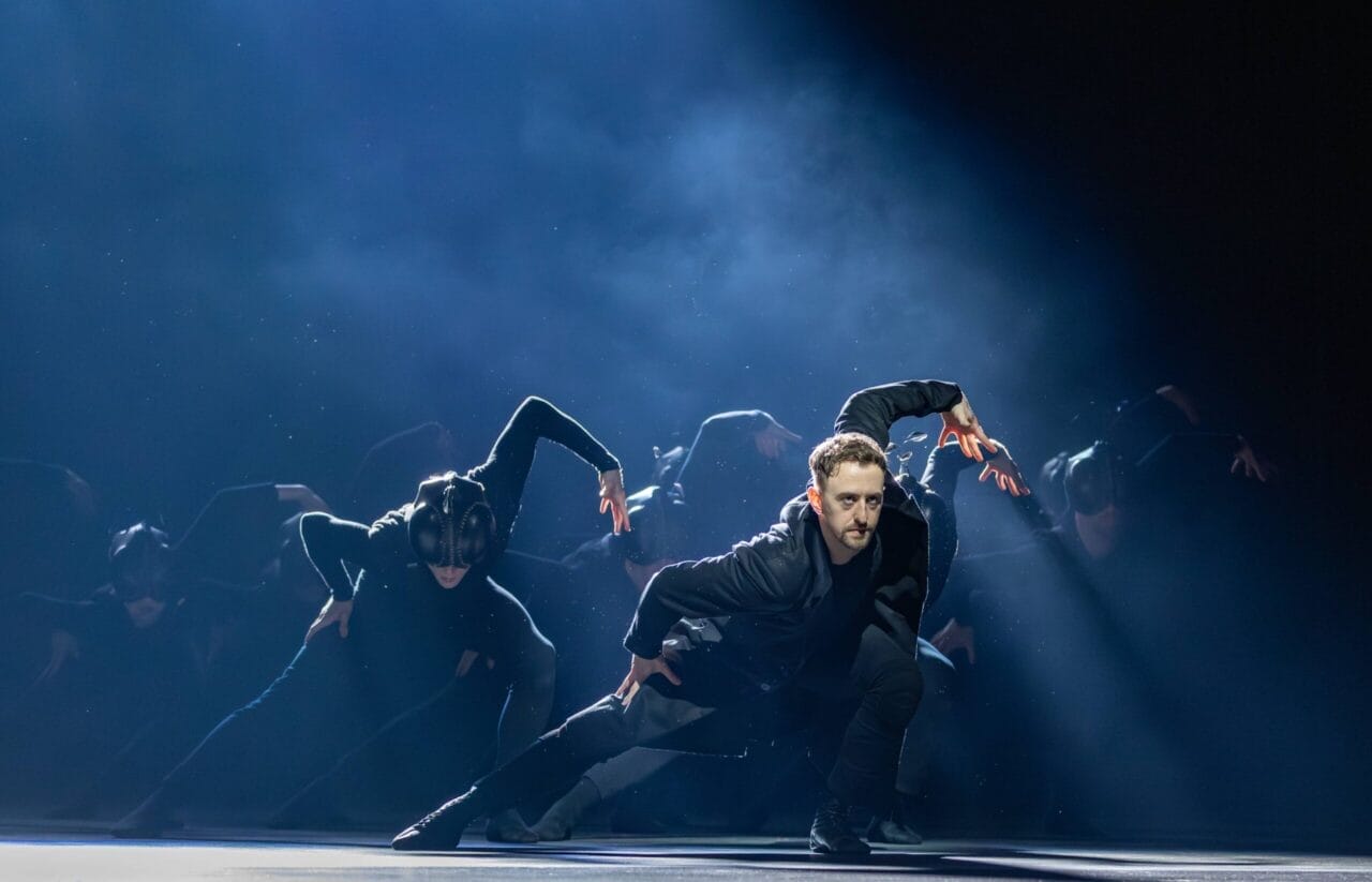 A group of dancers in black outfits performs a synchronized routine on stage under dramatic lighting, evoking the mysterious elegance of Mary, Queen of Scots.