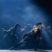 A group of dancers in black outfits performs a synchronized routine on stage under dramatic lighting, evoking the mysterious elegance of Mary, Queen of Scots.