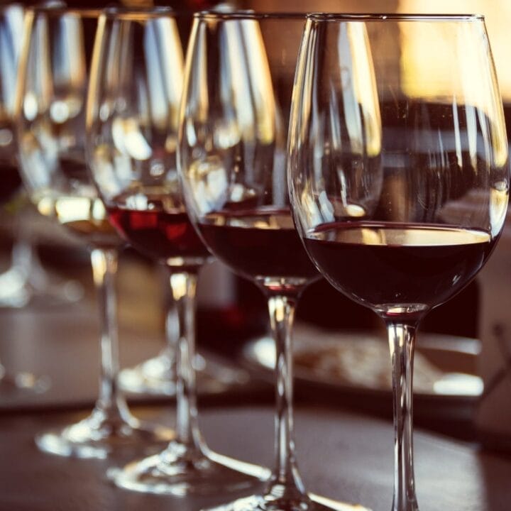 Several wine glasses filled with red and white wine are lined up on a table at a wine tasting, with sunlight reflecting off the glass surfaces.