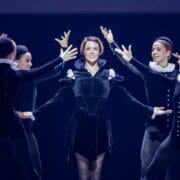 Five ballet dancers in black Renaissance-style costumes evoke Mary Queen of Scots on stage, with one dancer centered and others posing around her with outstretched arms, capturing a moment inspired by Scottish history.