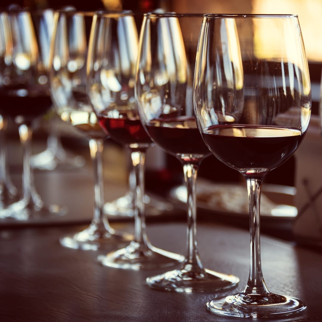 Five wine glasses, each containing different wines or amounts, are lined up on a table under a soft spotlight with a blurred background.