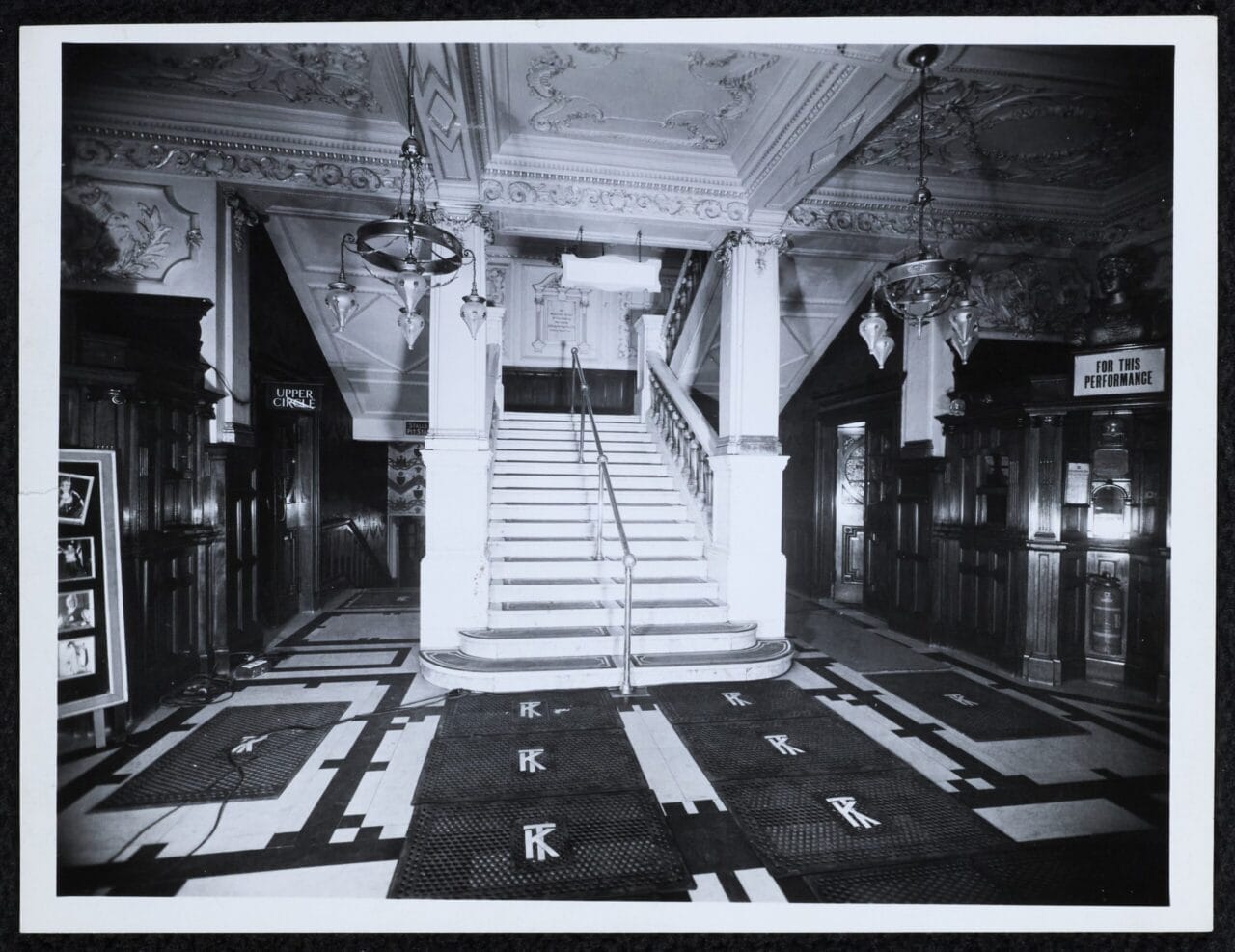 Black and white photo of The King's Theatre's ornate lobby with a grand staircase, decorative ceiling, chandeliers, and monogrammed floor mats.