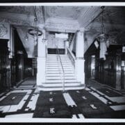 Black and white photo of The King's Theatre's ornate lobby with a grand staircase, decorative ceiling, chandeliers, and monogrammed floor mats.