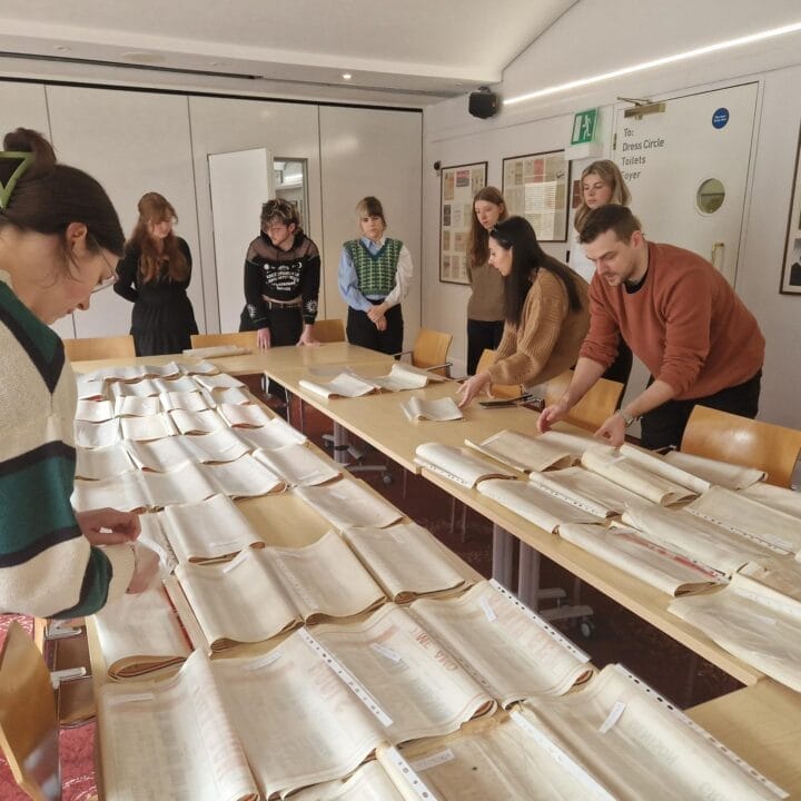 A group of people examines and organises large papers laid out on several tables in a brightly lit room.