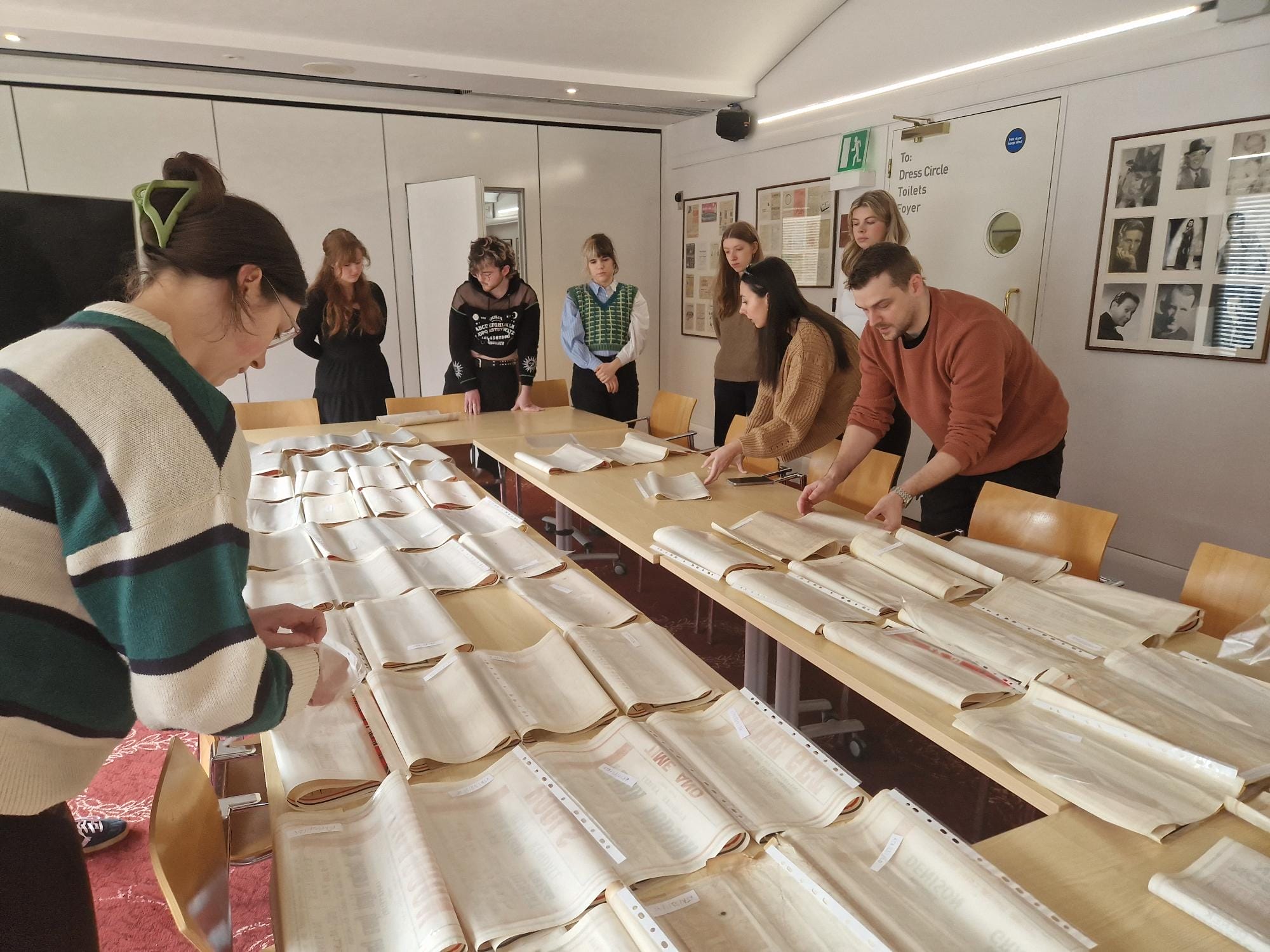 A group of people examines and organises large papers laid out on several tables in a brightly lit room.