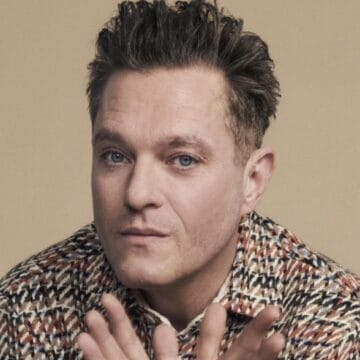 A man with short, dark hair wearing a patterned jacket looks directly at the camera with a neutral expression, hands raised in front of him.