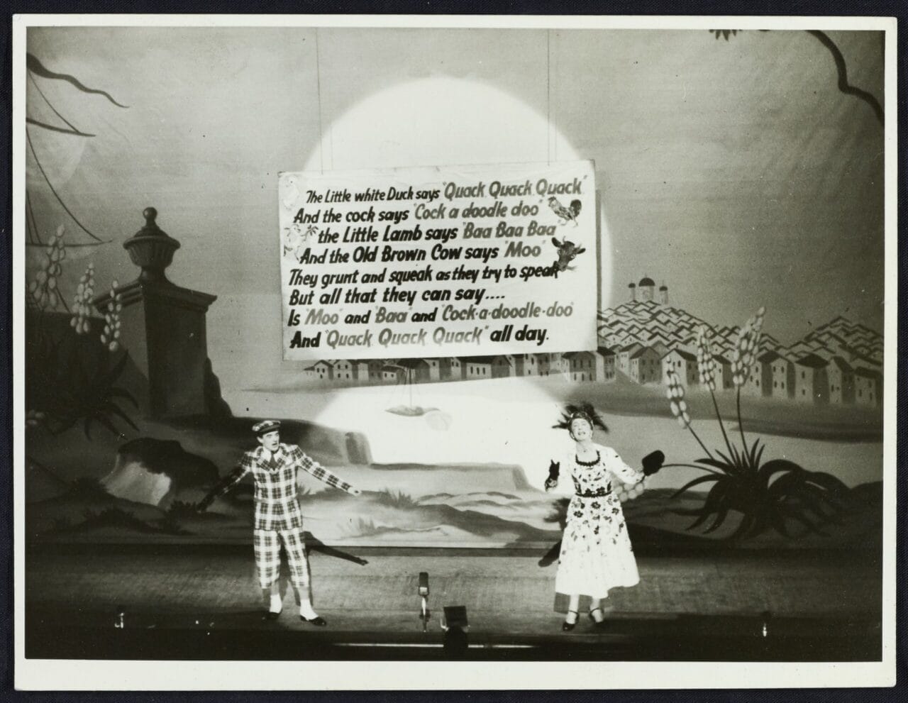 Two performers in costume stand on a stage with a rural backdrop, singing beneath a large sign displaying song lyrics about animal sounds.