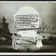Two performers in costume stand on a stage with a rural backdrop, singing beneath a large sign displaying song lyrics about animal sounds.