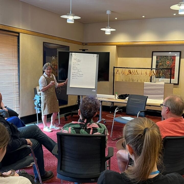 A woman stands by a flip chart leading a discussion with a seated group in a conference room.