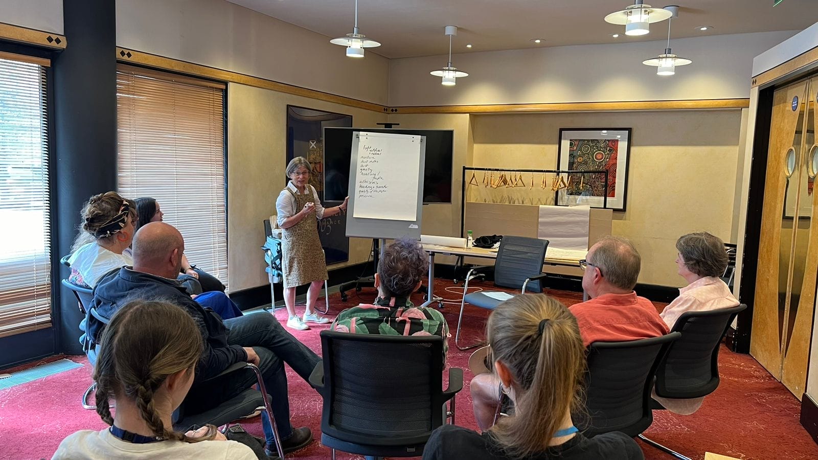 A woman stands by a flip chart leading a discussion with a seated group in a conference room.