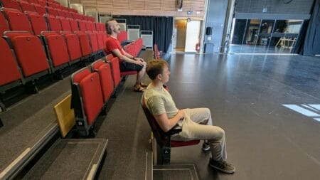 Two people sit apart in red theatre seats facing an empty stage in the spacious, well-lit Studio Theatre, enjoying easy access to the grand auditorium.