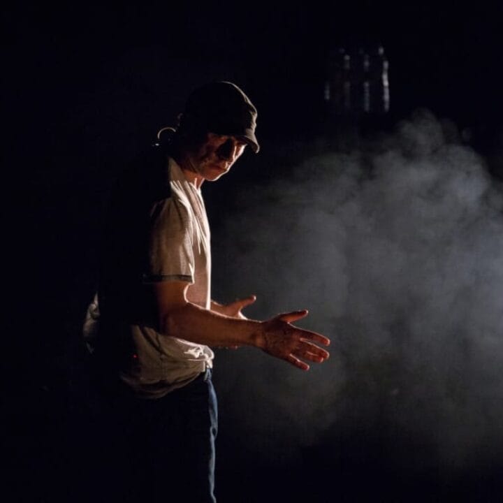 A man wearing a cap and t-shirt stands in a dark, smoky environment, illuminated by a single light source from the side.