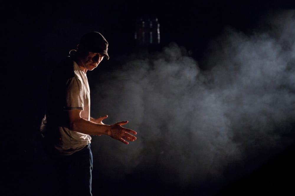 A man wearing a cap and t-shirt stands in a dark, smoky environment, illuminated by a single light source from the side.