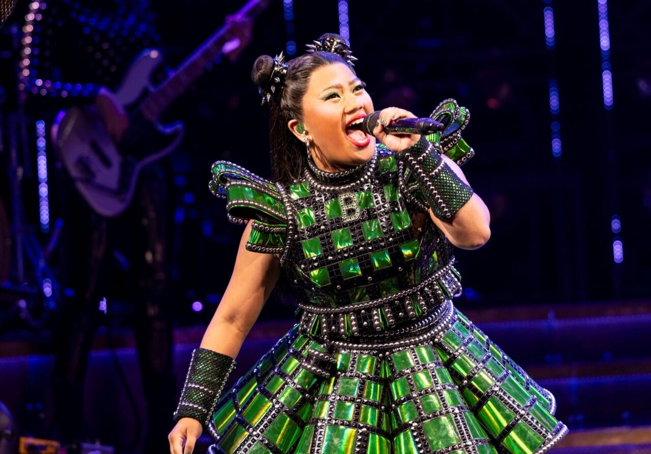 A performer wearing a shiny green costume sings into a microphone on stage, with a musician playing a guitar in the background.