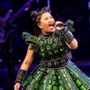A performer wearing a shiny green costume sings into a microphone on stage, with a musician playing a guitar in the background.