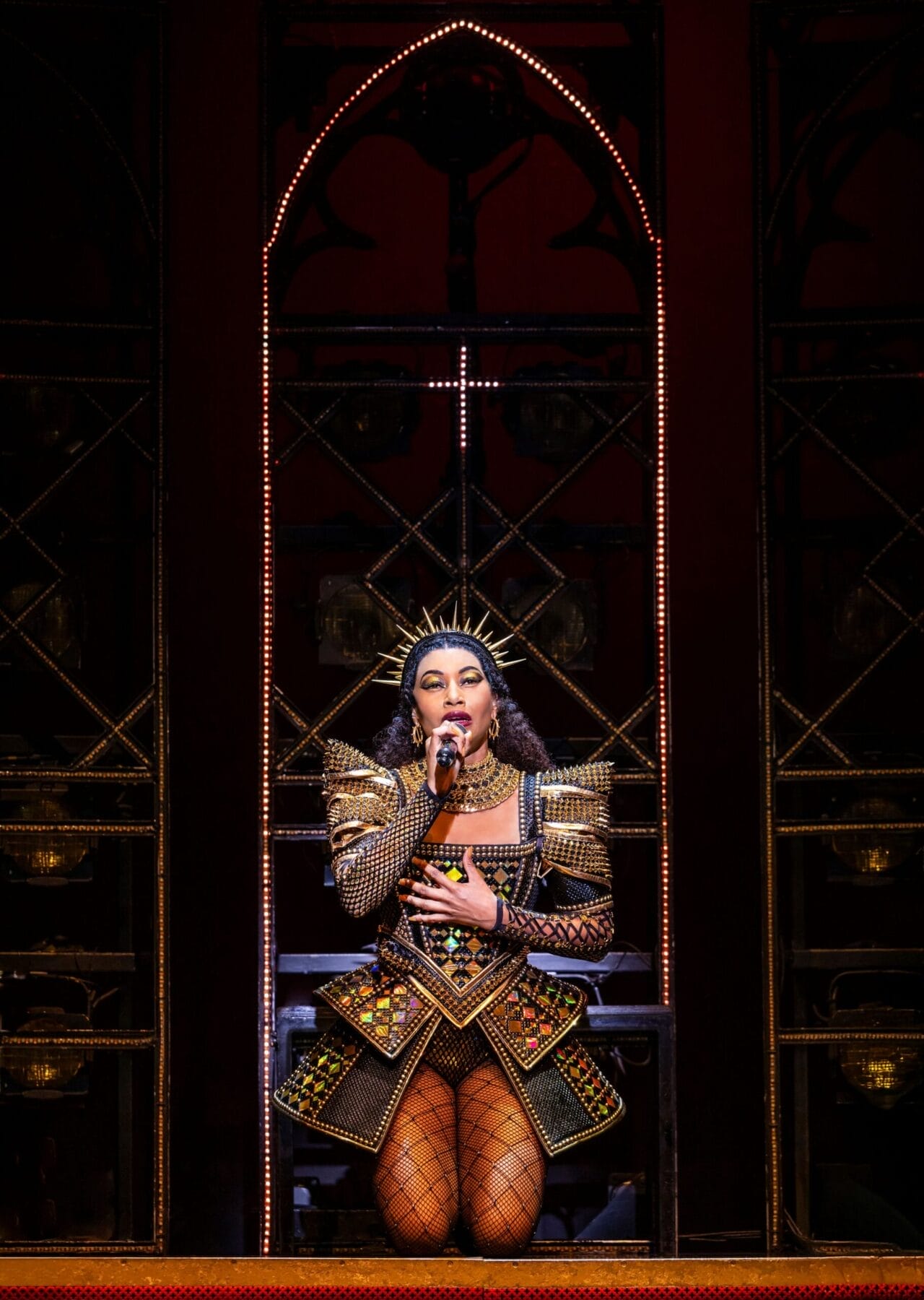 Performer in an ornate, gold and black costume with fishnet tights sings into a microphone on a stage with illuminated arches and dark background.