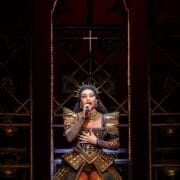 Performer in an ornate, gold and black costume with fishnet tights sings into a microphone on a stage with illuminated arches and dark background.