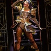 Performer in an ornate, gold and black, jewel-studded costume sings passionately on stage, holding a microphone and wearing a spiked crown.