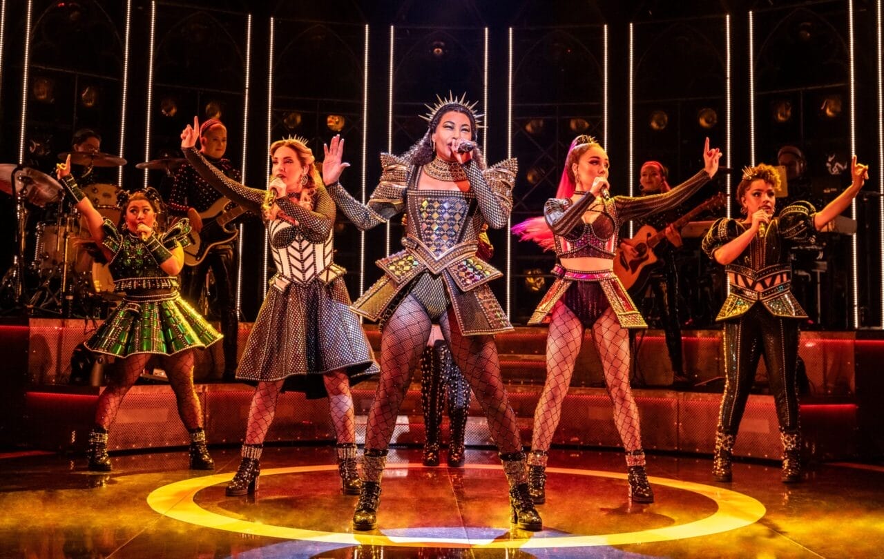 Five women in elaborate, metallic costumes perform energetically on stage with a live band in the background and dramatic lighting.