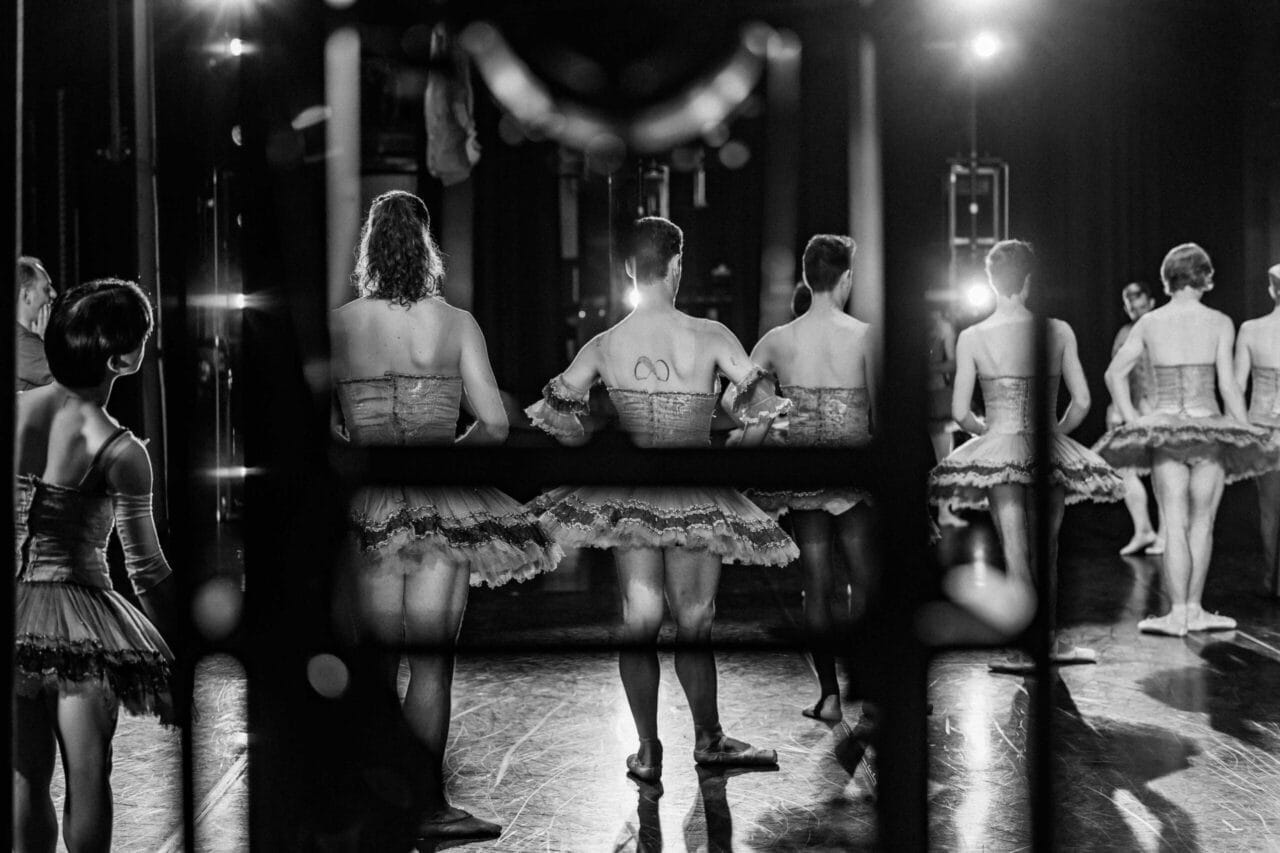 A black and white photo shows Les Ballets Trockadero dancers in tutus, viewed from behind through a window. They stand in a circle on stage, preparing to perform under bright ballet lights.