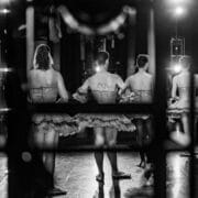 A black and white photo shows Les Ballets Trockadero dancers in tutus, viewed from behind through a window. They stand in a circle on stage, preparing to perform under bright ballet lights.