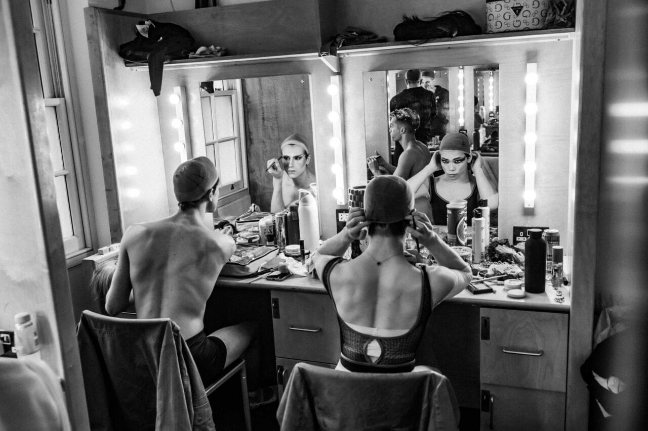 Two people sit at a make-up table, applying make-up and adjusting headwear in front of a mirror with lights. Various cosmetics and personal items are spread across the counter in a backstage Les Ballets Trockadero dressing room.