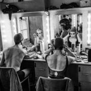 Two people sit at a make-up table, applying make-up and adjusting headwear in front of a mirror with lights. Various cosmetics and personal items are spread across the counter in a backstage Les Ballets Trockadero dressing room.