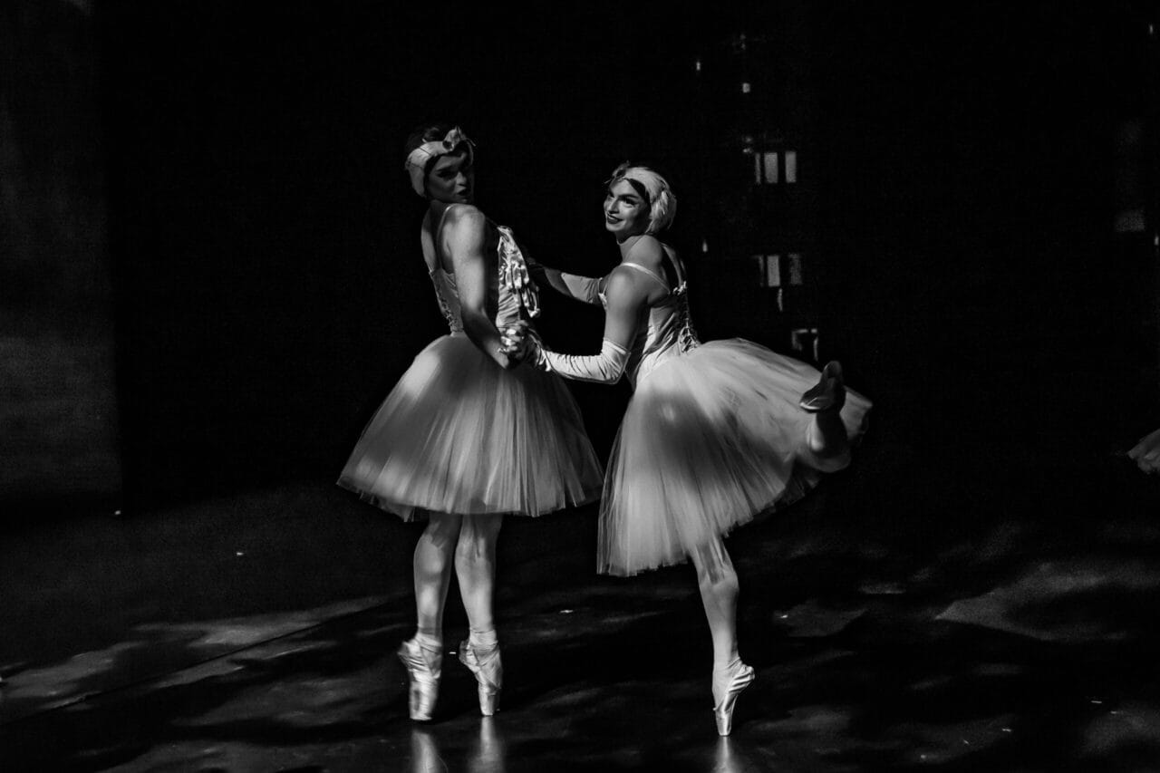Two Ballets Trockadero dancers in tutus and headpieces pose backstage. One stands en pointe with both feet, whilst the other balances en pointe with one leg bent behind her. Both face the camera in this dimly lit ballet scene reminiscent of Monte Carlo.