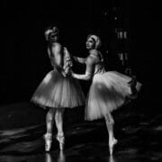 Two Ballets Trockadero dancers in tutus and headpieces pose backstage. One stands en pointe with both feet, whilst the other balances en pointe with one leg bent behind her. Both face the camera in this dimly lit ballet scene reminiscent of Monte Carlo.