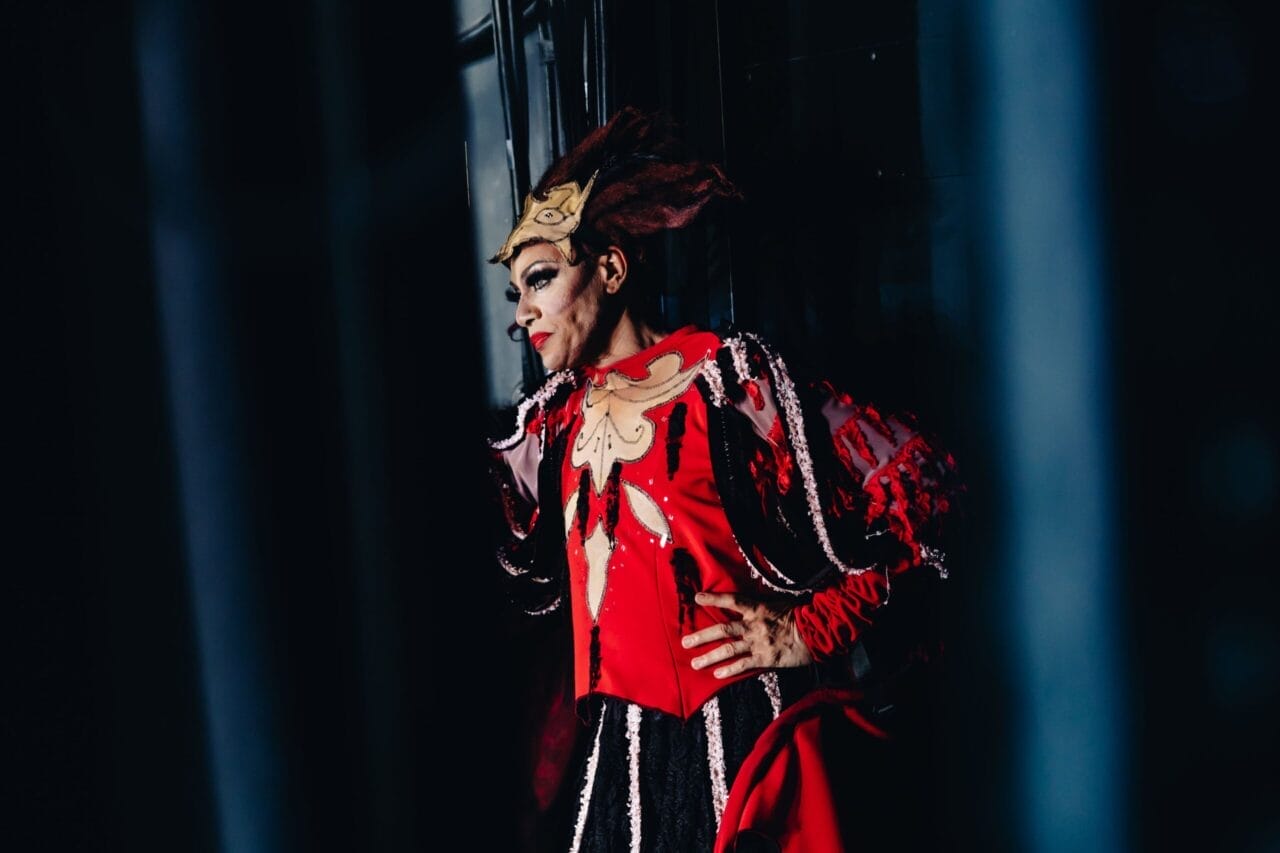A person in a bright red, elaborate costume with gold and brown mask-like headpiece stands against a dark background, posing with one hand on their hip and looking to the side, evoking the theatrical flair of Les Ballets Trockadero.