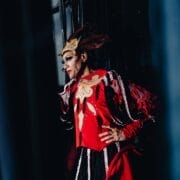 A person in a bright red, elaborate costume with gold and brown mask-like headpiece stands against a dark background, posing with one hand on their hip and looking to the side, evoking the theatrical flair of Les Ballets Trockadero.