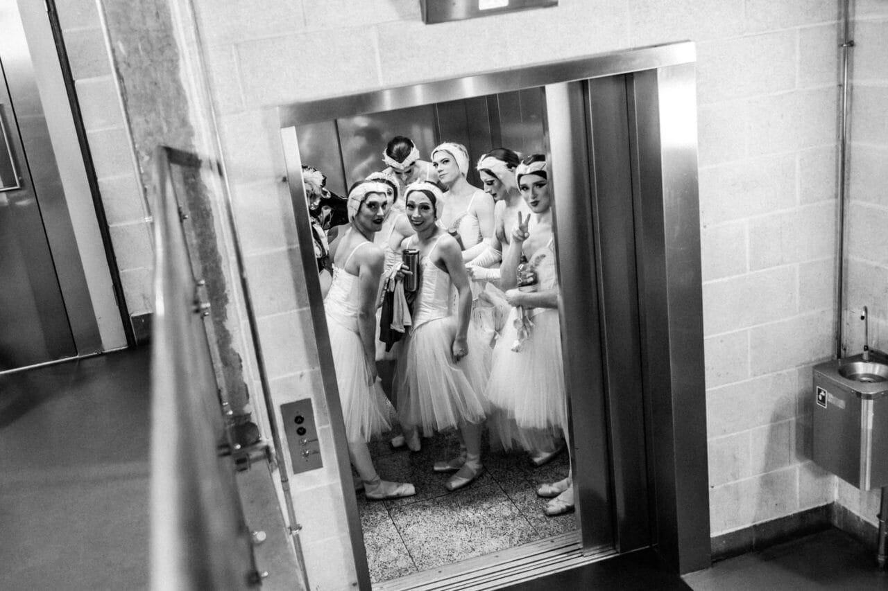 A group of ballet dancers in white costumes and headpieces from Les Ballets Trockadero stand inside an open lift, looking towards the camera in a backstage area with tiled walls and a metal rubbish bin.