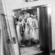 A group of ballet dancers in white costumes and headpieces from Les Ballets Trockadero stand inside an open lift, looking towards the camera in a backstage area with tiled walls and a metal rubbish bin.