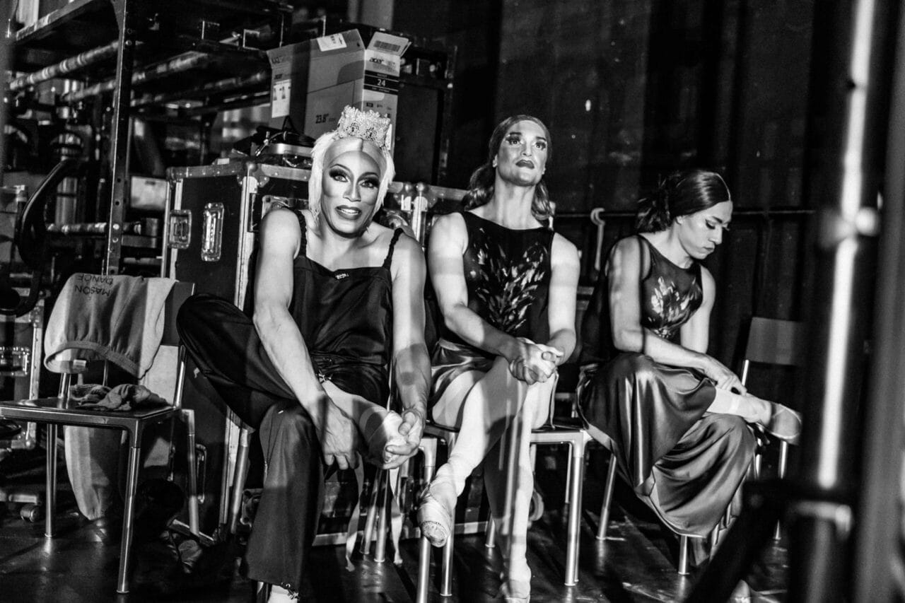 Three ballet dancers in costume sit backstage, surrounded by equipment and storage boxes at a Monte Carlo performance. Two adjust their pointe shoes while the third looks down. The black-and-white image captures the spirit of Les Ballets Trockadero.