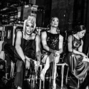 Three ballet dancers in costume sit backstage, surrounded by equipment and storage boxes at a Monte Carlo performance. Two adjust their pointe shoes while the third looks down. The black-and-white image captures the spirit of Les Ballets Trockadero.