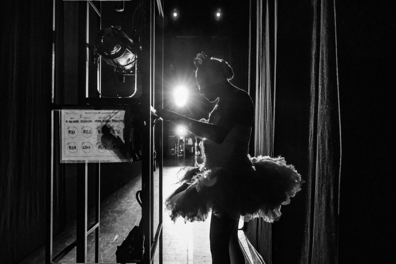 A ballerina in a tutu stands backstage in silhouette, illuminated by bright stage lights, with stage equipment and curtains visible around her—a quiet moment before the ballet performance at Monte Carlo.