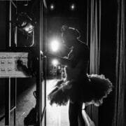 A ballerina in a tutu stands backstage in silhouette, illuminated by bright stage lights, with stage equipment and curtains visible around her—a quiet moment before the ballet performance at Monte Carlo.